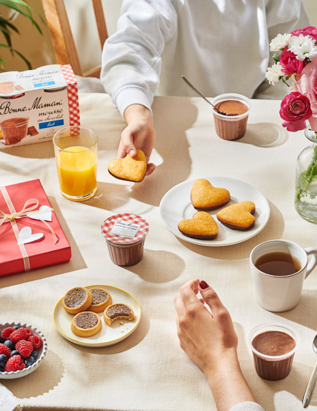 Photographie culinaire d’un goûter gourmand avec des produits Bonne Maman tels que des gâteaux en forme de cœur, des mousses au chocolat, du jus d’orange et une tasse de thé, disposés sur une table lumineuse pour une ambiance conviviale.