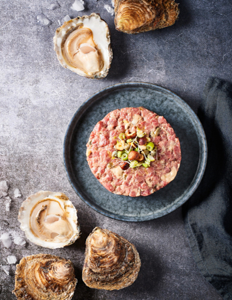 Photographie culinaire d’un tartare de bœuf et huîtres plates La Perle de Marie-Jo, garni de câpres, noisettes torréfiées et jeunes pousses, entouré de coquilles d’huîtres sur une table en pierre grise.