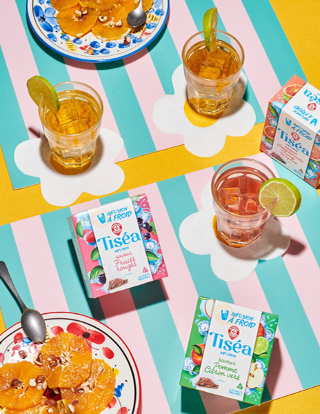 Photographie culinaire mettant en scène les infusions à froid Tiséa de Leclerc avec des verres de thé glacé, des rondelles de citron vert et des assiettes d’oranges aux amandes, sur une table colorée à rayures pastel.
